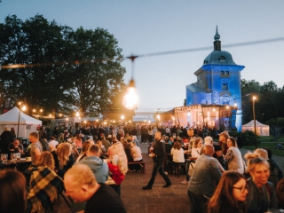 Valmiermuižas etnomūzikas festivāla vakara atmosfēra – apmeklētāji pulcējušies pie skatuves un galdiem, fonā izgaismota baznīca un gaismu virtenes.