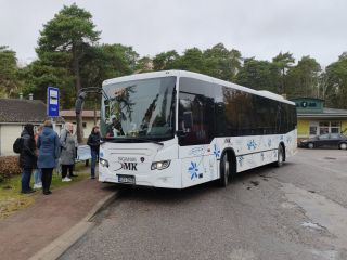 Balts autobuss pieturā Ainažos uzņem pasažierus. Pie autobusa durvīm stāv vairāki cilvēki, bet fonā redzami koki, ēkas un stāvlaukums. Uz pieturas zīmes ir norāde “Ainaži”, bet autobusa maršruta displejā redzams numurs 741.