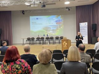 Konferences zāle ar sēdvietām auditorijai. Uz skatuves pie tribīnes runātājs uzstājas pasākumā par Igaunijas un Latvijas pārrobežu sabiedriskā transporta savienojumu atjaunošanu. Aiz runātāja redzams prezentācijas ekrāns ar projekta ROCC informāciju un va