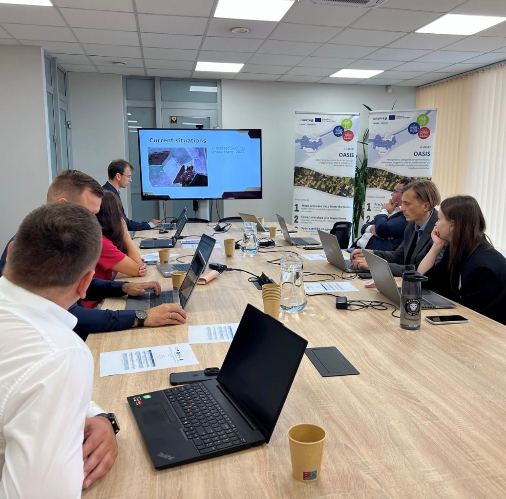 Projekta partneru sanāksme konferenču telpā. Dalībnieki sēž pie garā galda ar klēpjdatoriem un klausās prezentāciju uz ekrāna, kurā redzami satelītattēlu dati par lauksaimniecības zemēm. Telpā izvietoti projekta OASIS informatīvie stendi, bet uz galda atrodas dokumenti, ūdens krūkas un vienreizlietojamās krūzes. Attēls ilustrē starptautisku sadarbību un digitālo risinājumu izmantošanu lauksaimniecības datu analīzē.