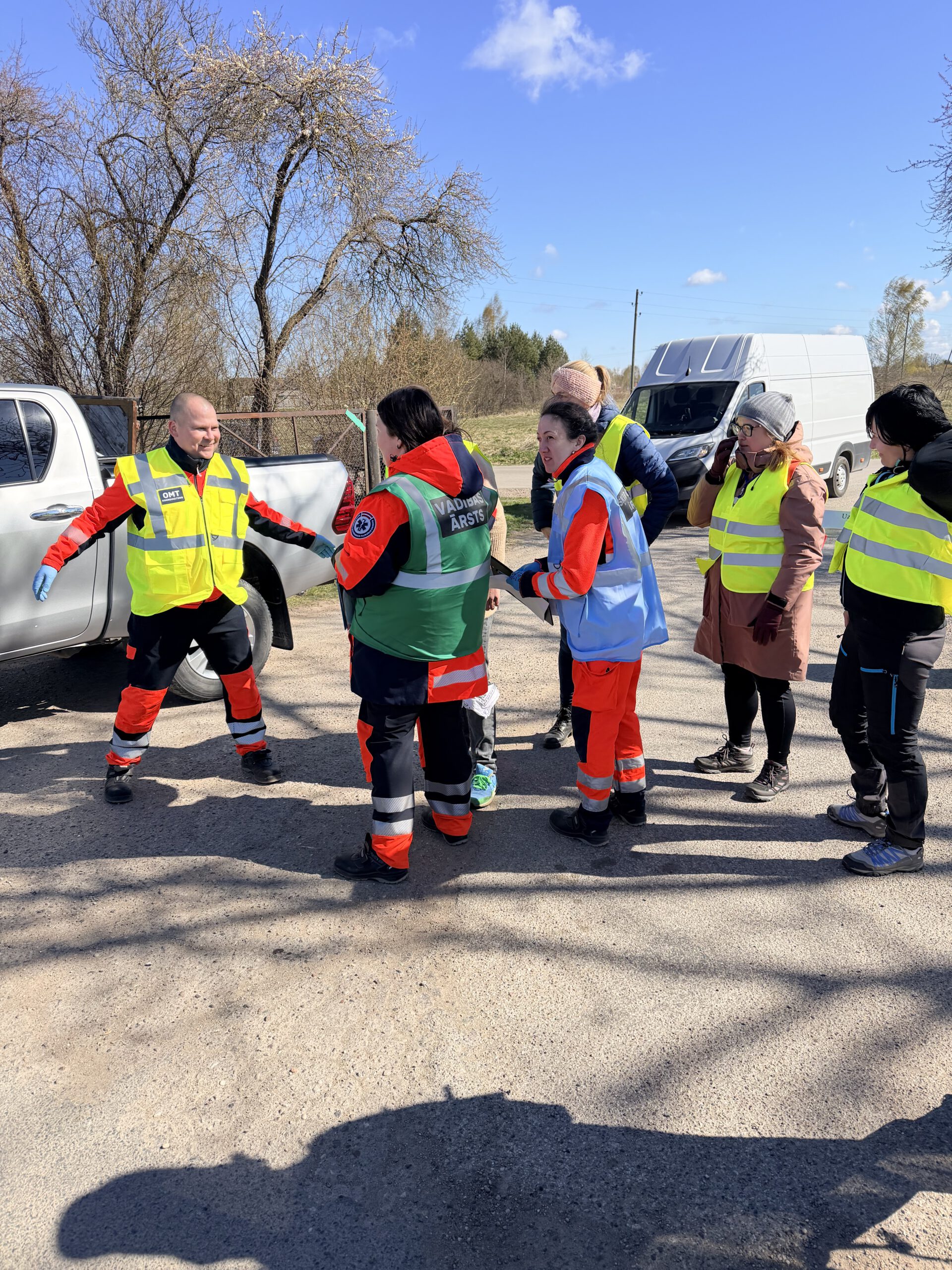 Brīvprātīgo un mediķu kopīgās mācības brīvā dabā: operatīvo dienestu darbinieki un brīvprātīgie atstarojošās vestēs apspriež uzdevumu, priekšplānā redzams mediķis ar uzrakstu "VADĪBAS ĀRSTS".
