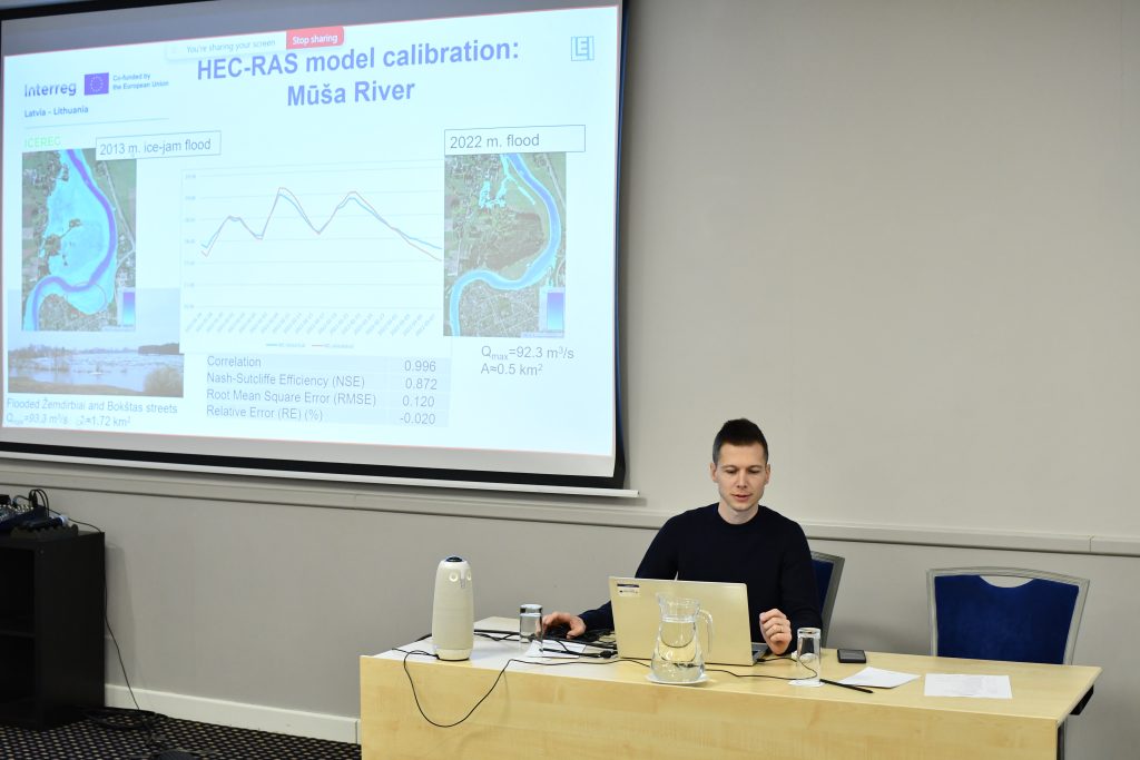 Vīrietis sēž pie galda ar klēpjdatoru un vada tiešsaistes prezentāciju, kurā redzams "Interreg Latvija–Lietuva" slaids par HEC-RAS modeļa kalibrēšanu Mūsas upei (Mūša River), salīdzinot 2013. un 2022. gada plūdu datus.