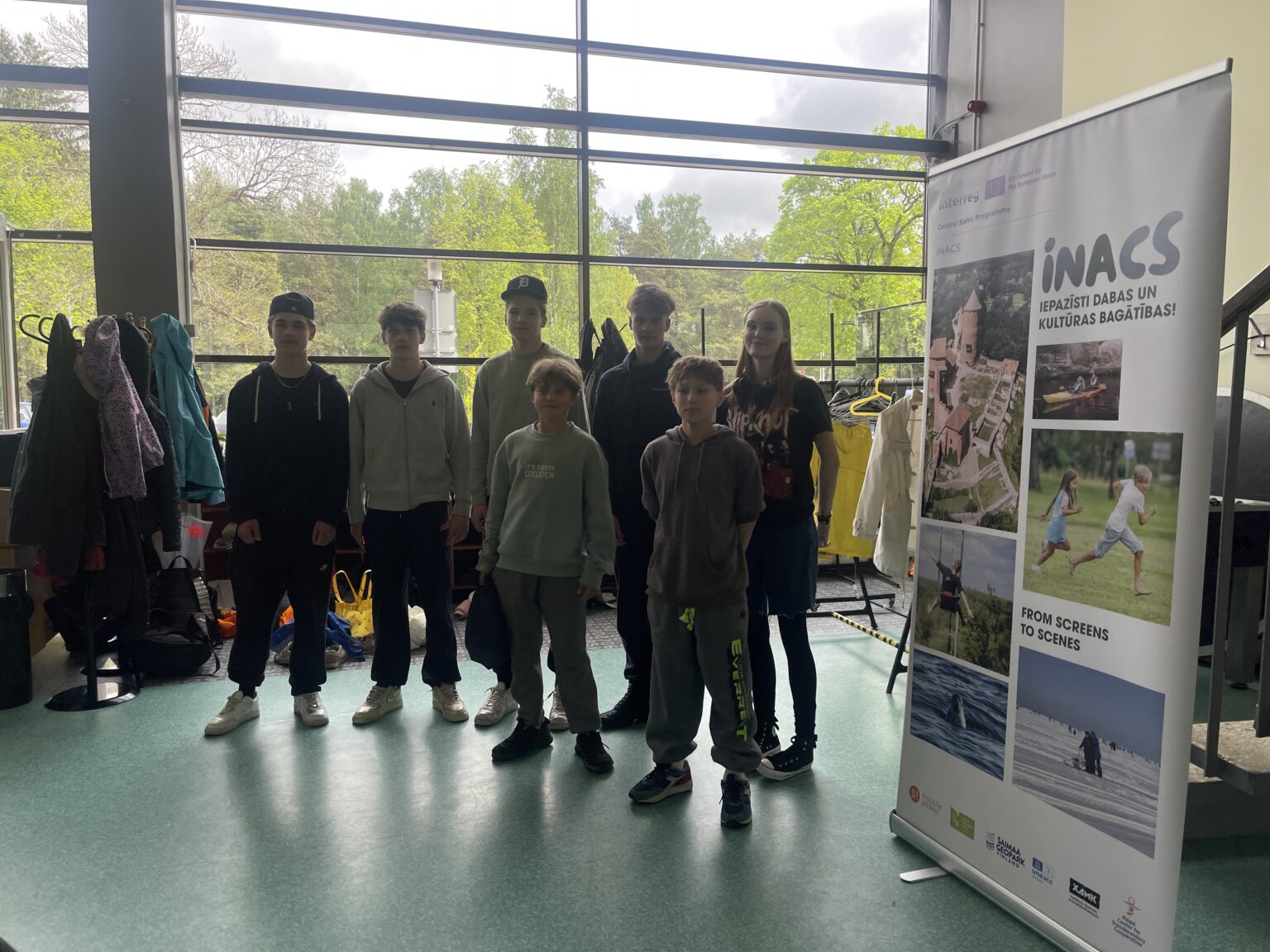 Septiņi jaunieši nostājušies blakus projekta INACS stendam ar uzrakstu “Iepazīsti dabas un kultūras bagātības!” un dabas un kultūrvēsturisko vietu fotogrāfijām.